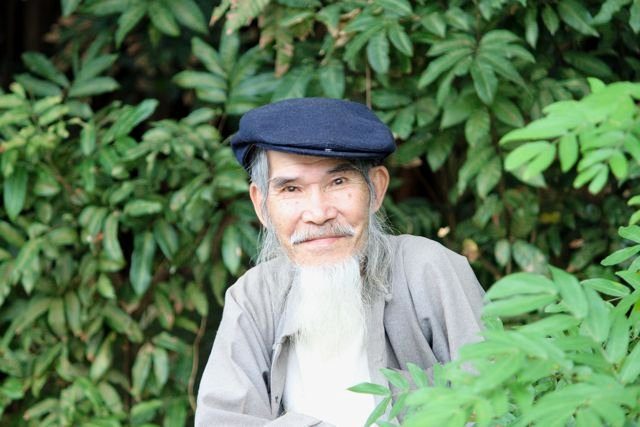 Elderly man with a long white beard and flat cap among green foliage