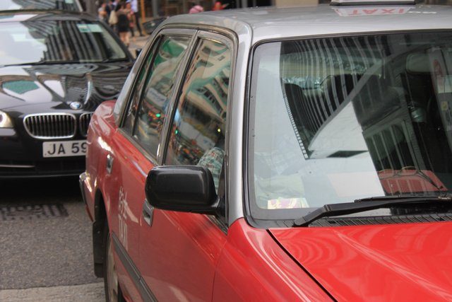 Red Hong Kong taxi in traffic, city buildings reflected in its windows